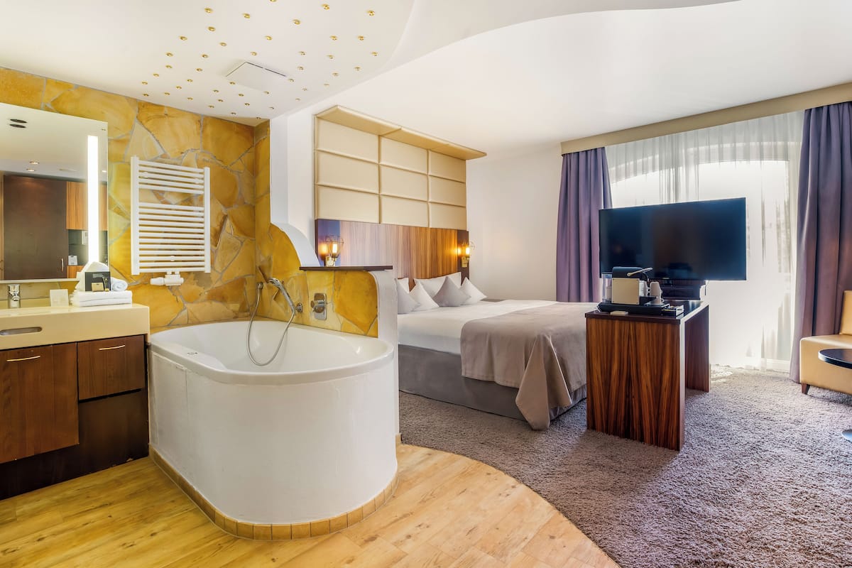 A modern hotel room with a large bathtub, a bed, a television, and a wooden floor.