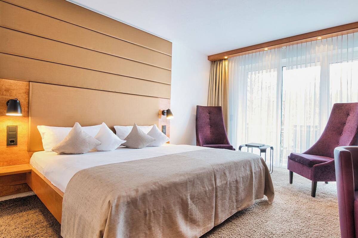 A hotel room with a large bed, two chairs, a nightstand, and a window with curtains.