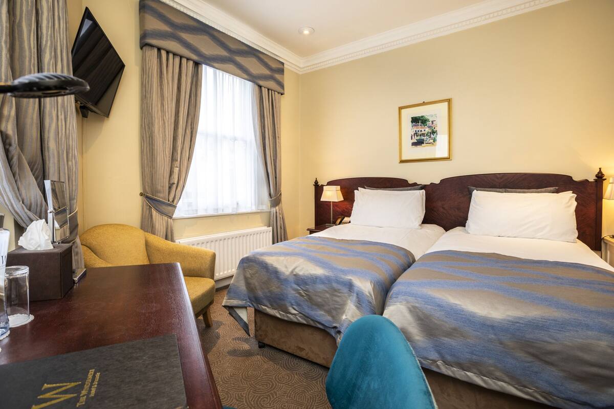 A hotel room with two beds, a desk, a chair, a TV mounted on the wall, and a window with curtains.