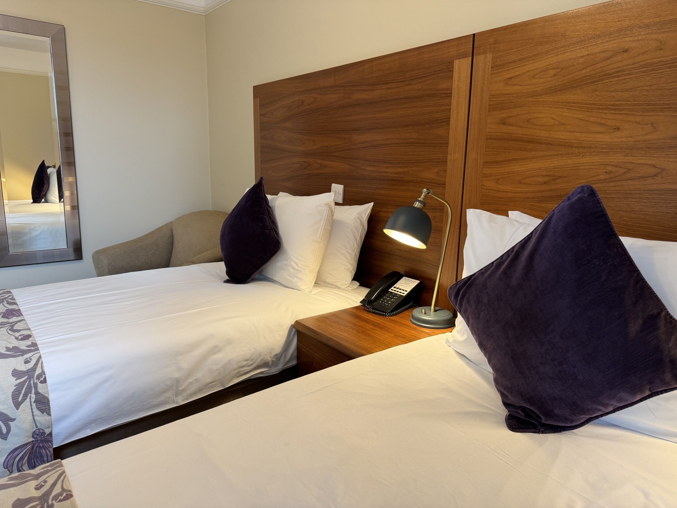 A hotel room with two beds, a wooden headboard, a bedside table with a lamp, and a telephone.