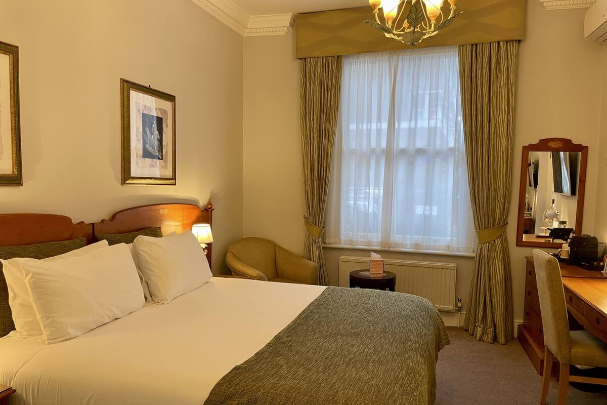 A hotel room with a large bed, a desk, a chair, a lamp, a window with curtains, and a mirror.