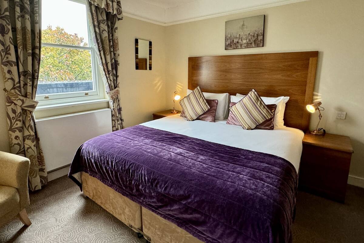 A neatly arranged bedroom with a large bed, two bedside tables with lamps, a window with curtains, and a cityscape painting on the wall.
