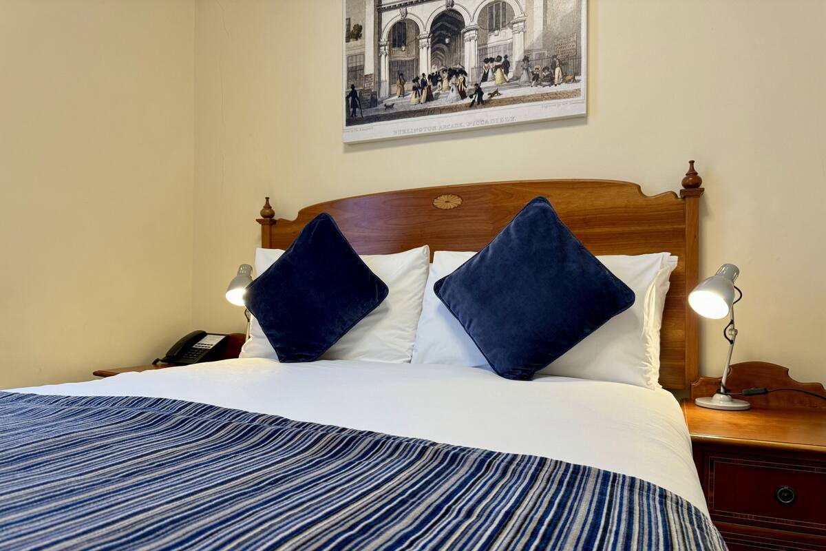 A neatly made bed with a striped bedspread, two dark pillows, and a wooden headboard. There is a nightstand with a lamp and a telephone. A framed picture hangs on the wall above the bed.