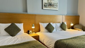 A hotel room with two beds, each with a wooden headboard, white bedding, and green throw pillows. There is a nightstand with a lamp and a phone on each bed. A framed picture hangs on the wall above the beds. - The Windermere Hotel, London (London)