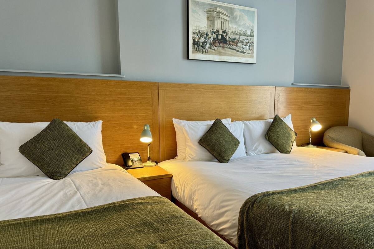 A hotel room with two beds, each with a wooden headboard, white bedding, and green throw pillows. There is a nightstand with a lamp and a phone on each bed. A framed picture hangs on the wall above the beds.