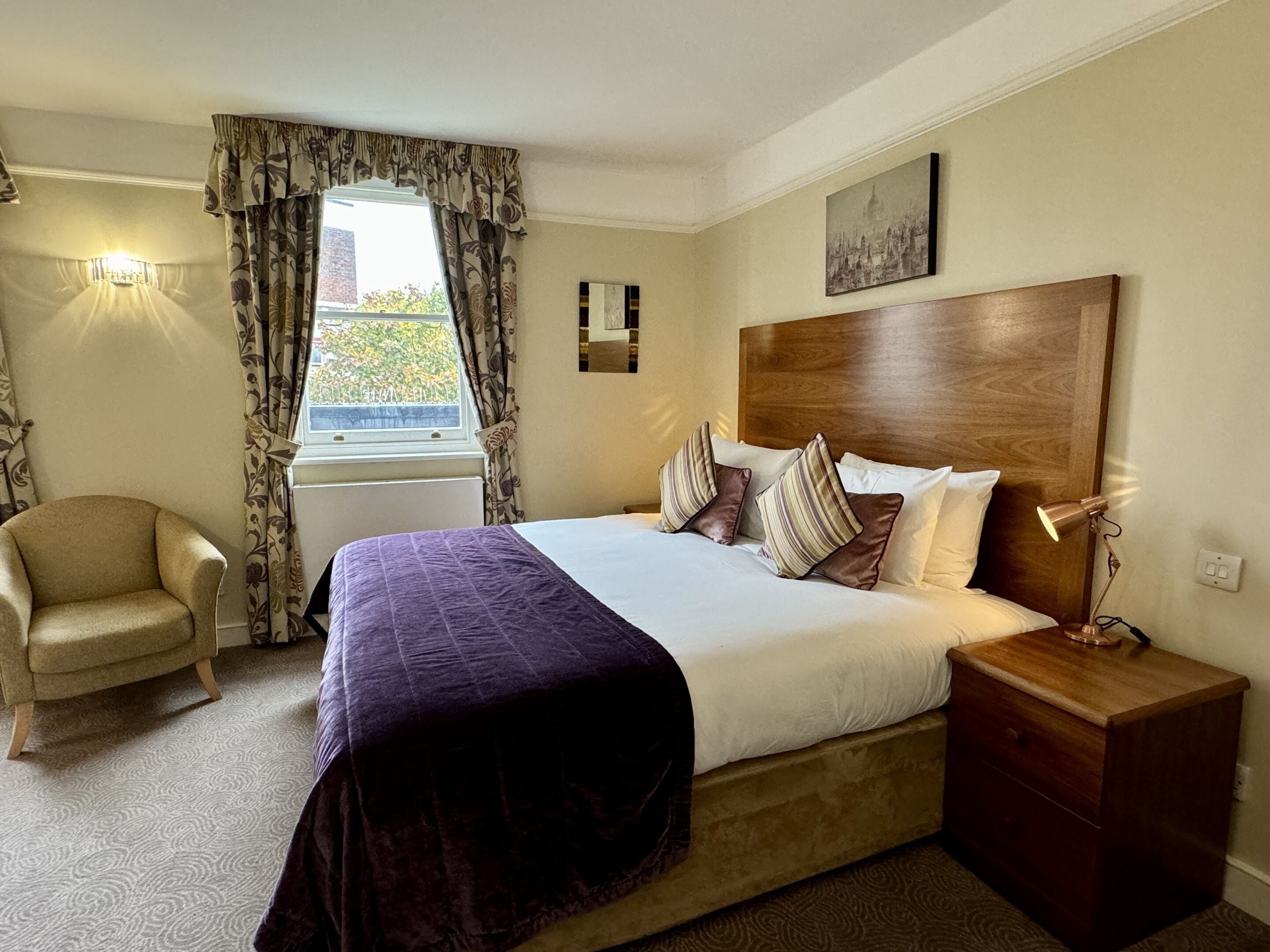 A hotel room with a bed, a chair, a nightstand, a lamp, a window with curtains, and a painting on the wall.