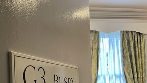 A hotel room with a sign reading "G3 BUSBY" on the wall, a striped armchair, and a window with curtains. - The Windermere Hotel, London (London)