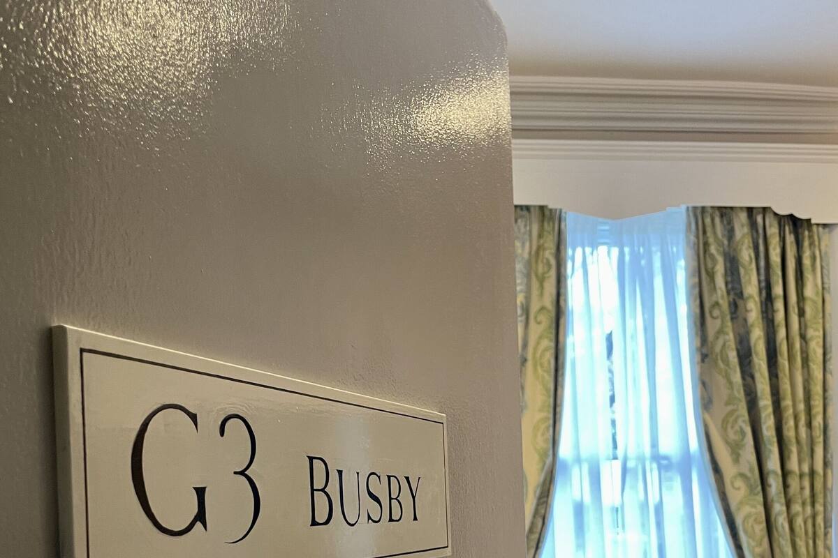 A hotel room with a sign reading "G3 BUSBY" on the wall, a striped armchair, and a window with curtains.