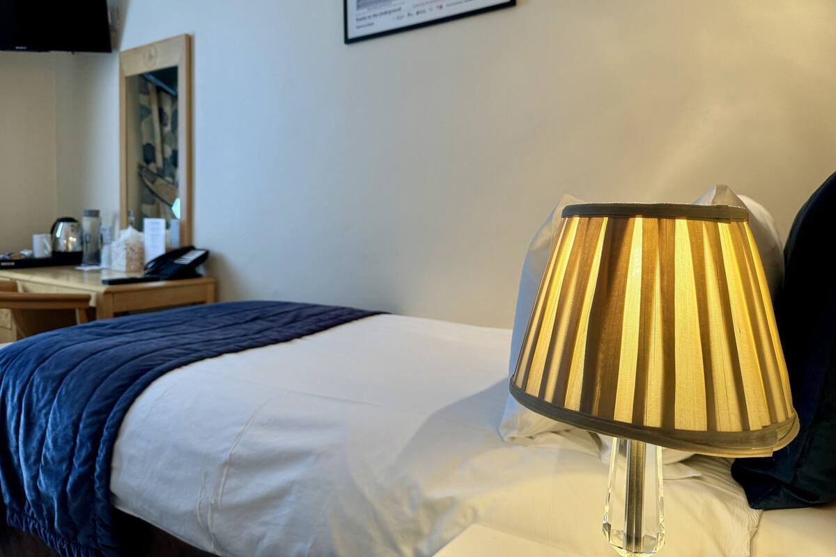 A hotel room with a bed, a bedside table, a lamp, a framed picture on the wall, and a sign with text.