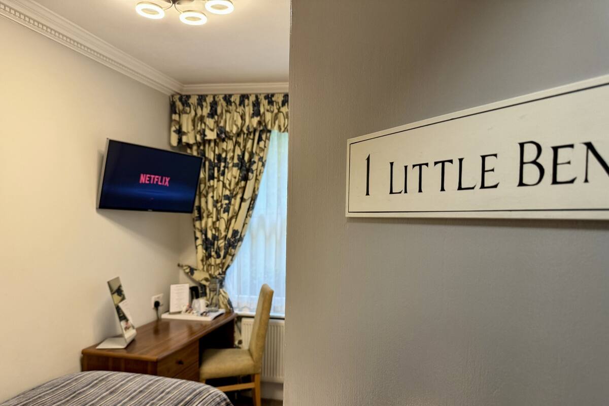 A hotel room with a bed, a desk, a chair, a TV displaying Netflix, and a sign that reads "I LITTLE BEN".