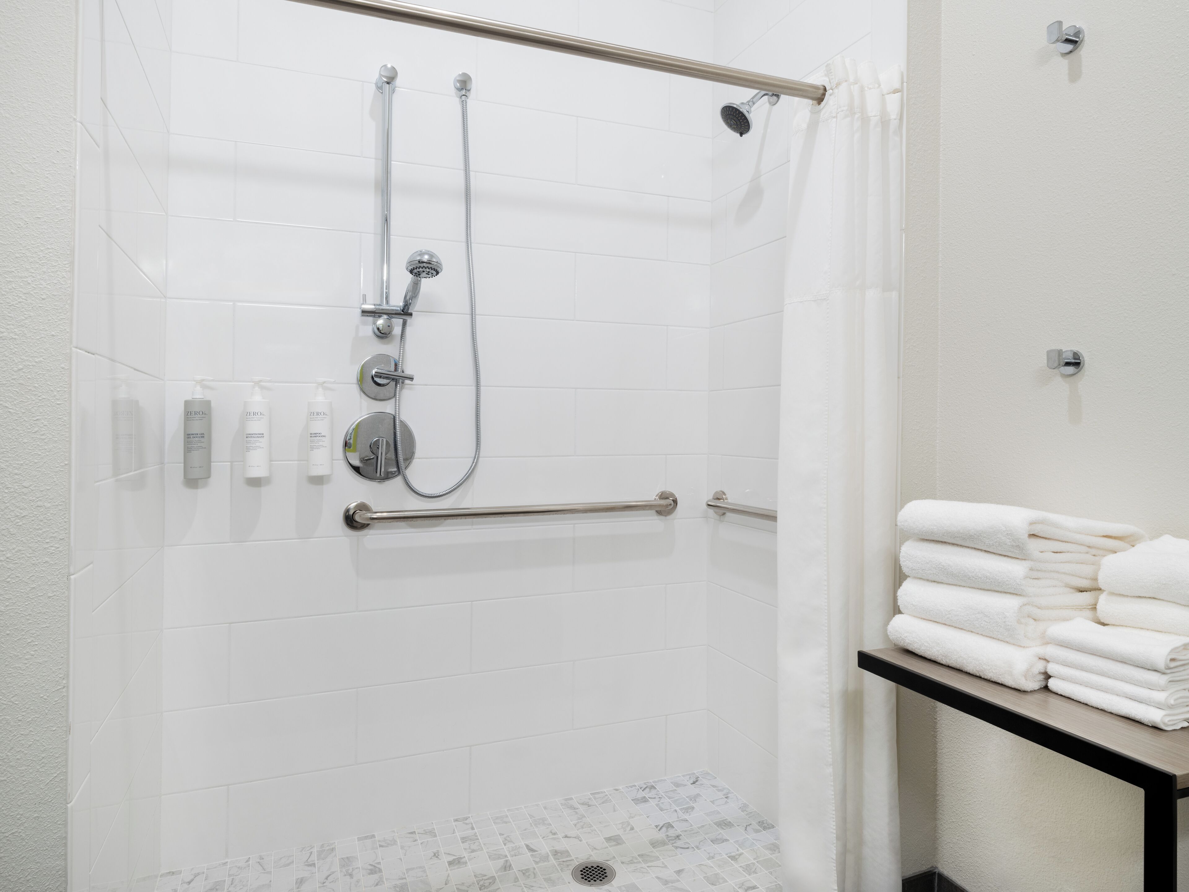 A shower with a handheld showerhead, a wall-mounted faucet, and a handheld showerhead. There are two grab bars installed for safety. A towel rack with folded towels is visible.