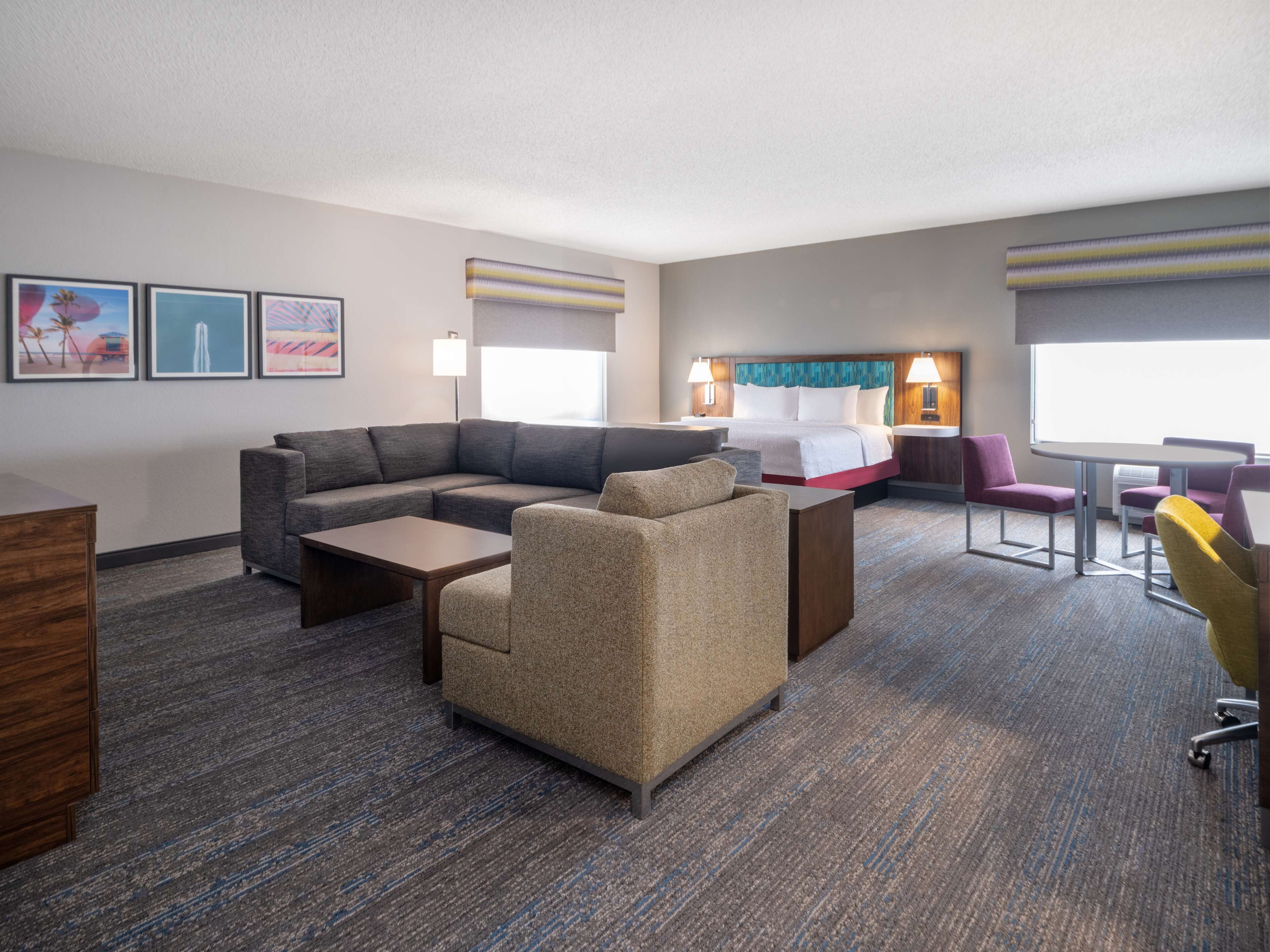 A hotel room with a sectional sofa, a small armchair, a desk, a bed with a headboard, and a dining table with chairs.