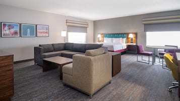 A hotel room with a sectional sofa, a small armchair, a desk, a bed with a headboard, and a dining table with chairs.