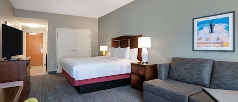 A hotel room with a bed, a TV, a desk, a chair, a couch, and a painting on the wall.