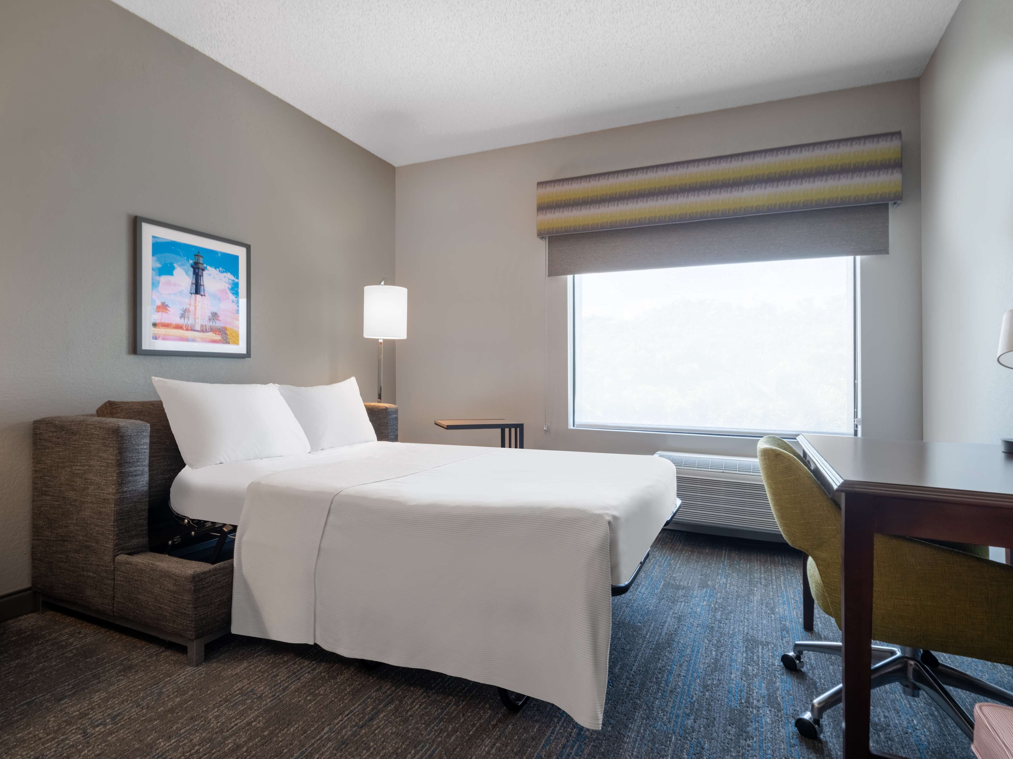 A hotel room with a bed, a desk, a chair, a lamp, a picture on the wall, and a window.