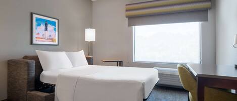 A hotel room with a bed, a desk, a chair, a lamp, a picture on the wall, and a window.