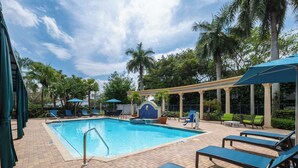 A swimming pool with lounge chairs and umbrellas, surrounded by palm trees and a covered area with columns. - Hampton Inn Boca Raton-Deerfield Beach (Deerfield Beach)