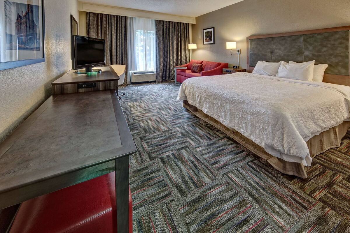A hotel room with a bed, a desk with a TV, a red chair, and a window with curtains.