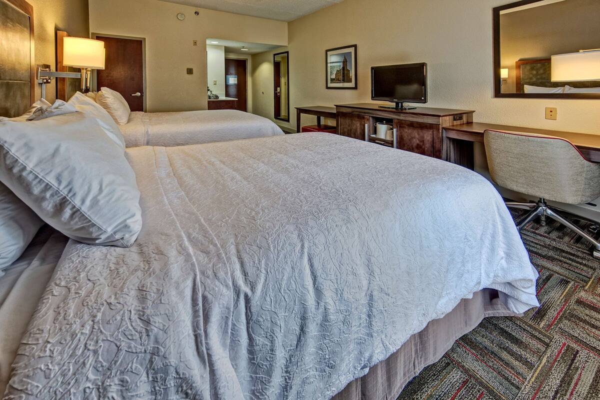 A hotel room with two beds, a desk, a chair, a television, and a mirror.