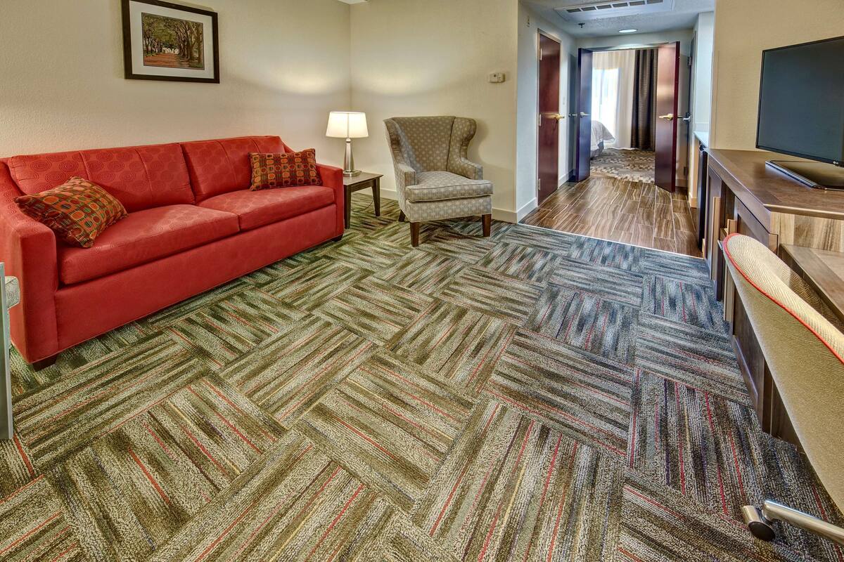 A hotel room with a red sofa, a patterned carpet, a chair, a TV, and a hallway leading to another room.