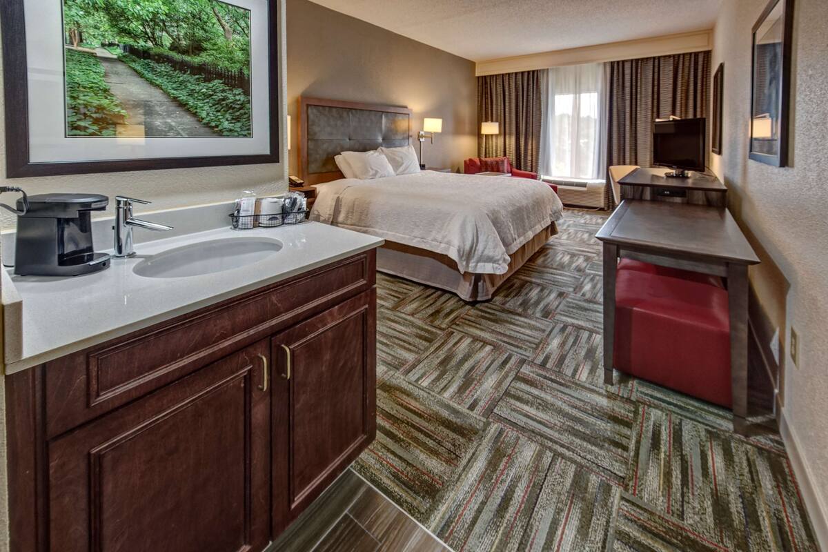 A hotel room with a large bed, a desk, a red chair, a TV, and a bathroom with a sink and mirror.