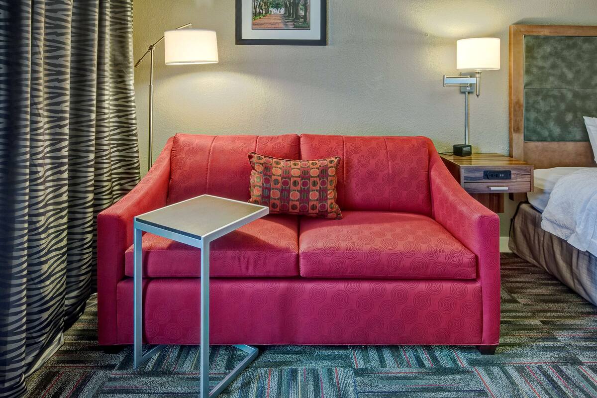 A red sofa with a patterned pillow, a small table, and a lamp on a nightstand.