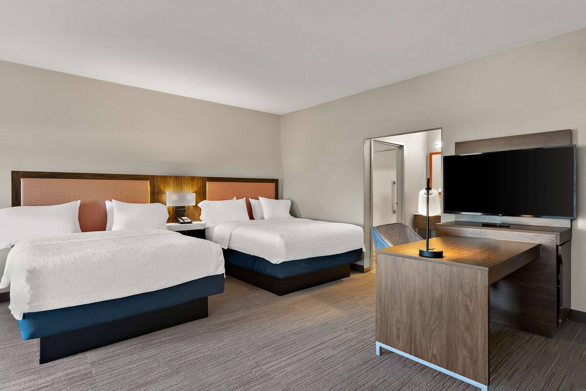 Quarto de hotel com duas camas, uma secretária e uma televisão, além de um espelho.