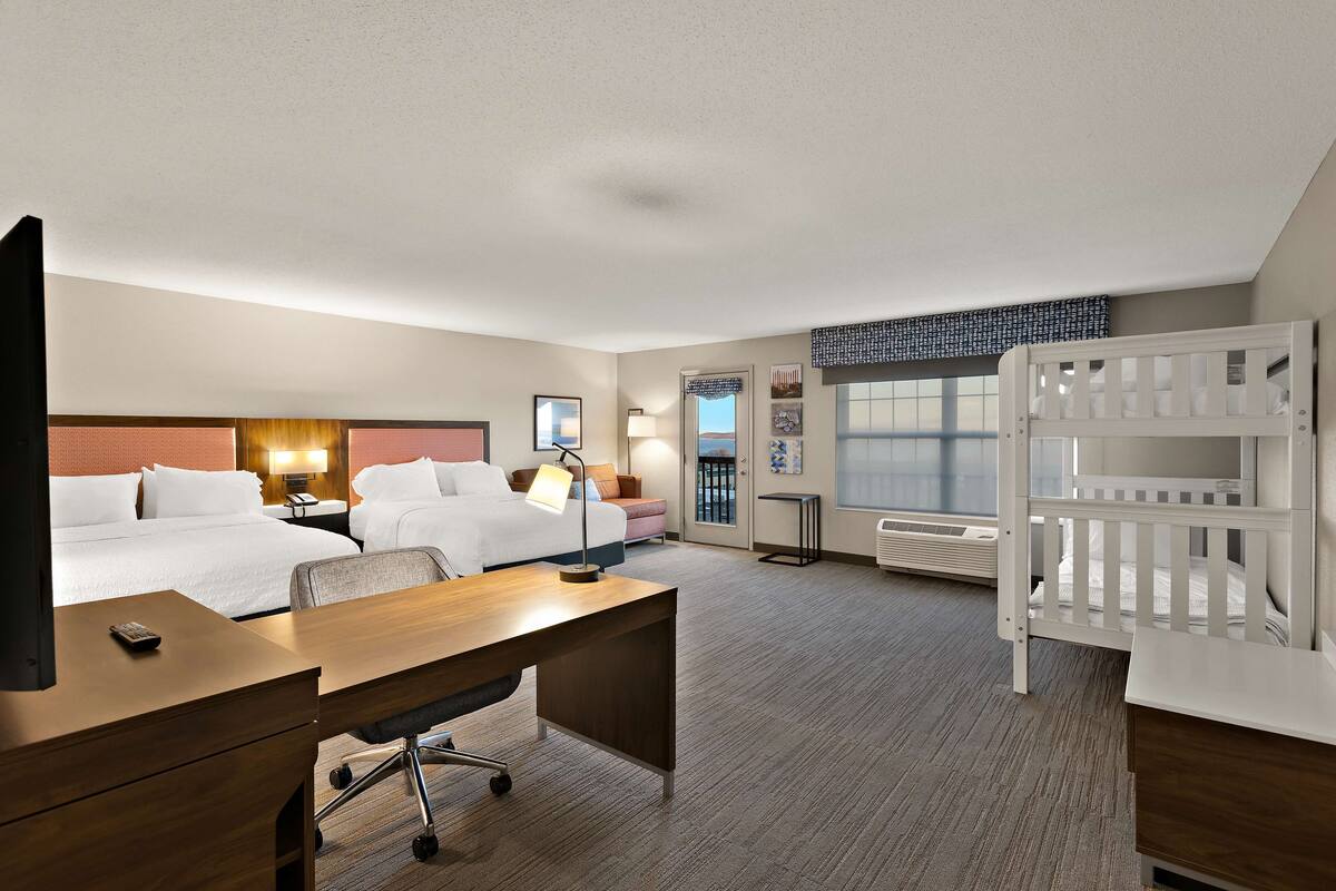 Quarto de hotel com escrivaninha, duas camas, berço, televisão, luminária e janela com vista.