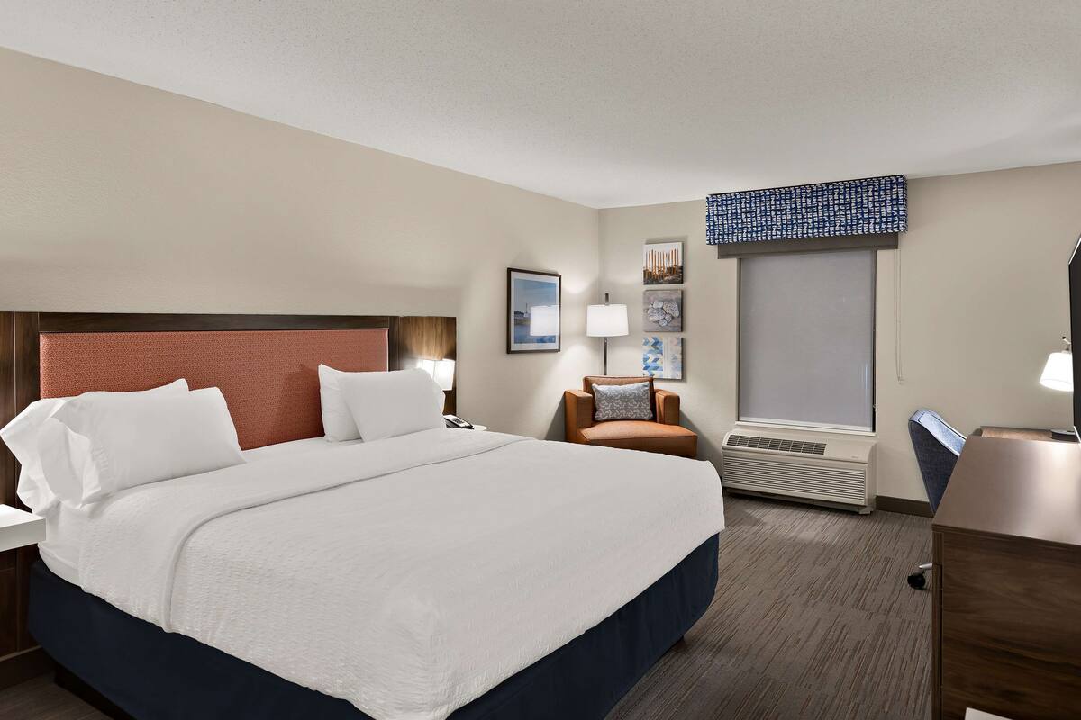 Quarto de hotel com cama grande, escrivaninha, cadeira, janela com persianas e televisão presa à parede.