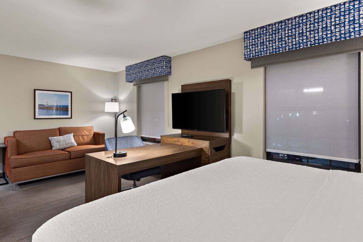 Quarto de hotel com cama, escrivaninha com luminária, televisão, sofá e janela com persianas.