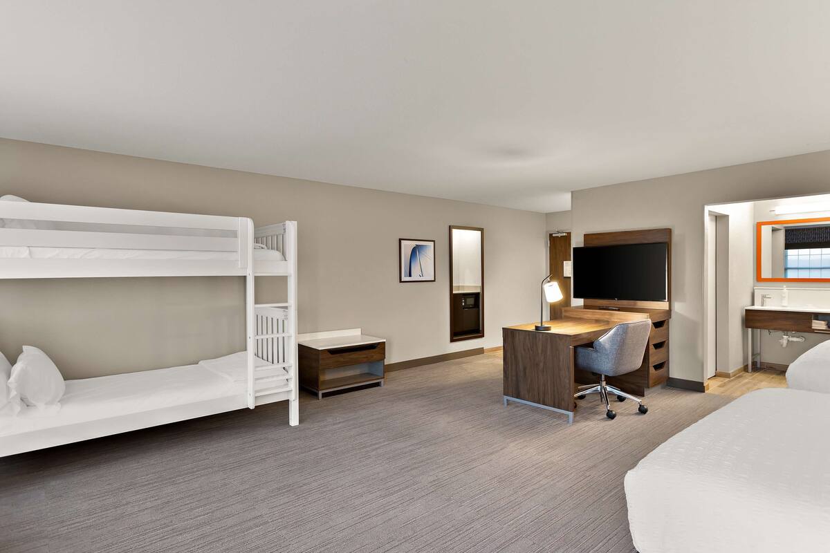 Quarto de hotel com beliches, uma escrivaninha, uma cadeira, uma televisão e uma cama.