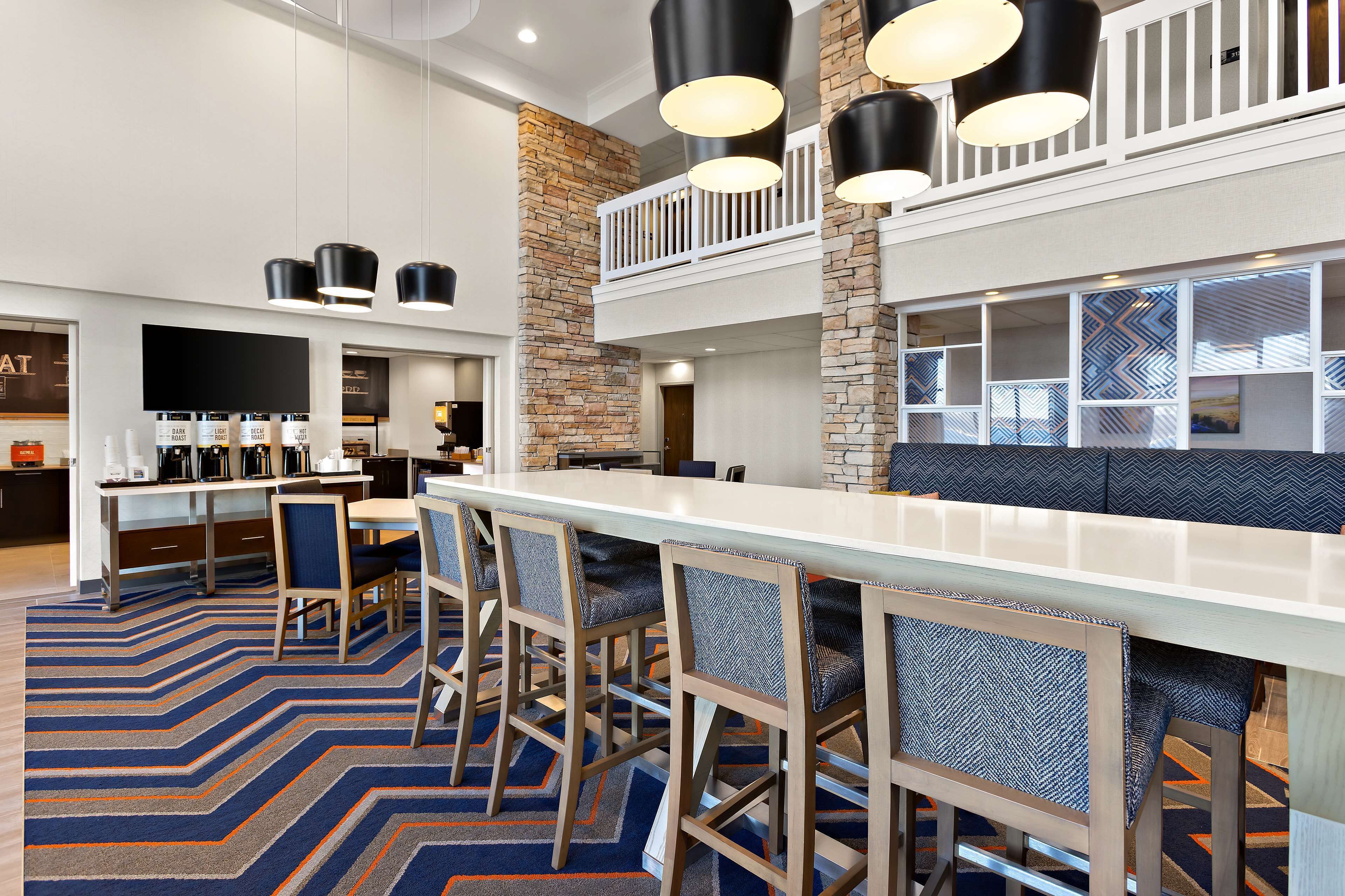 A modern hotel lobby with a long reception desk, bar area with high stools, and a seating area with a patterned rug.