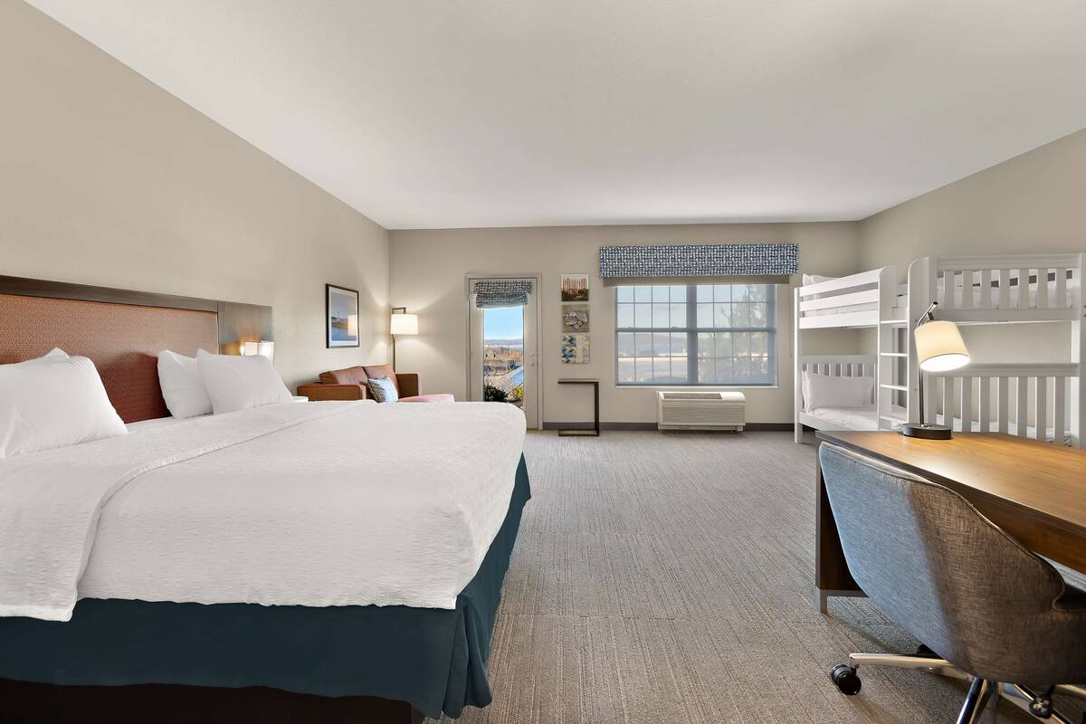 Quarto de hotel com uma cama grande, uma escrivaninha, uma cadeira, janela com persianas, radiador e vista para a cidade.