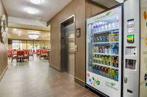 Un pasillo de hotel con una máquina expendedora llena de bebidas, un comedor con sillas rojas y un ascensor.