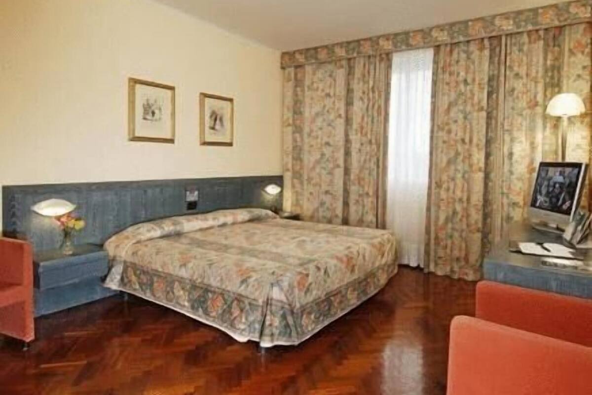 A hotel room with a bed, a desk with a lamp, a chair, and two framed pictures on the wall.