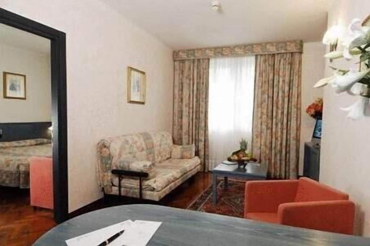 A room with a sofa, a small table with a pineapple, a chair, and a bed in the background.
