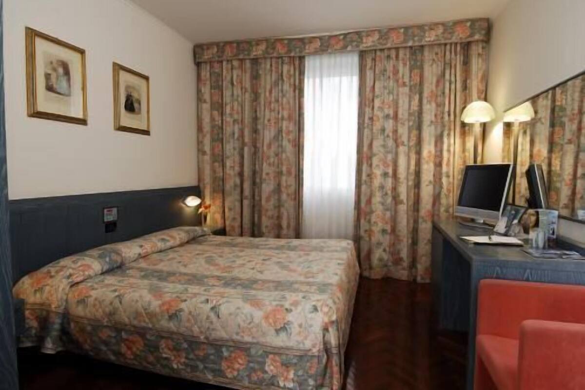 A hotel room with a bed, a desk with a computer, a chair, and two framed pictures on the wall.