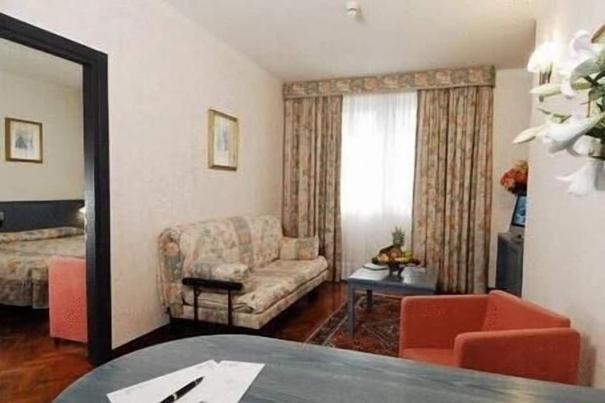 A compact hotel room with a sofa, a small table with a pineapple, a chair, and a bed in the adjacent room.