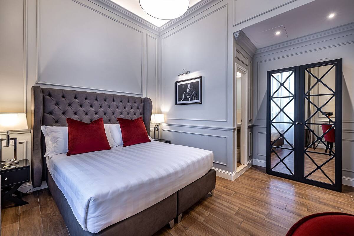 A bedroom with a tufted headboard, a bed with white and red bedding, a nightstand with a lamp, and a wooden floor.