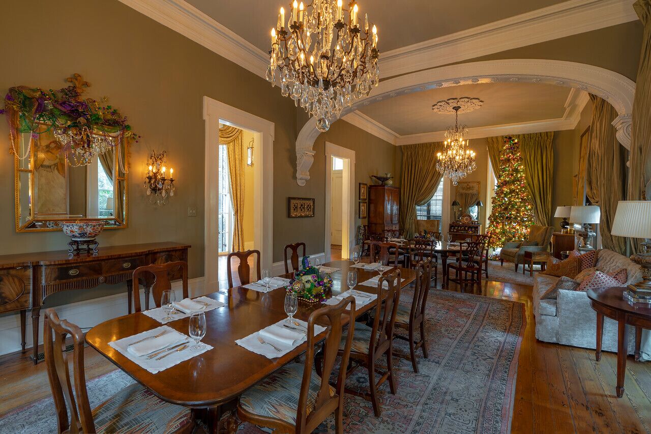 A formal dining room with a large wooden table set for six, surrounded by chairs. A chandelier hangs from the ceiling, and a Christmas tree is visible in the background.