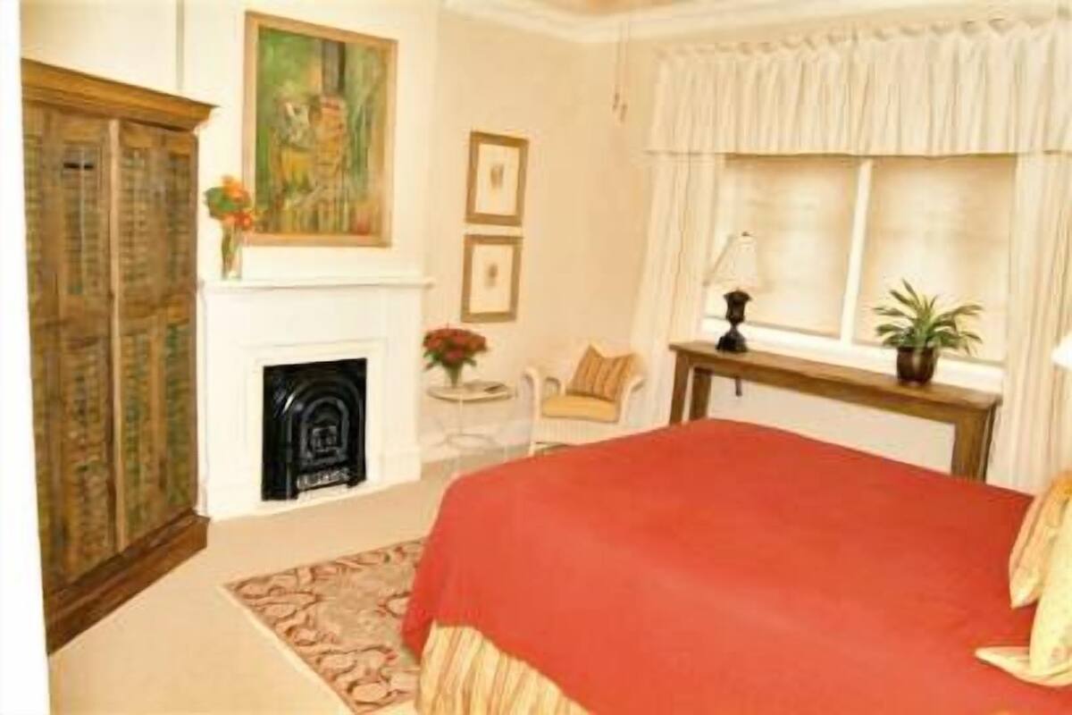 Quarto com cama grande, lareira, janela com cortinas, uma cadeira e mesa pequena com abajur.