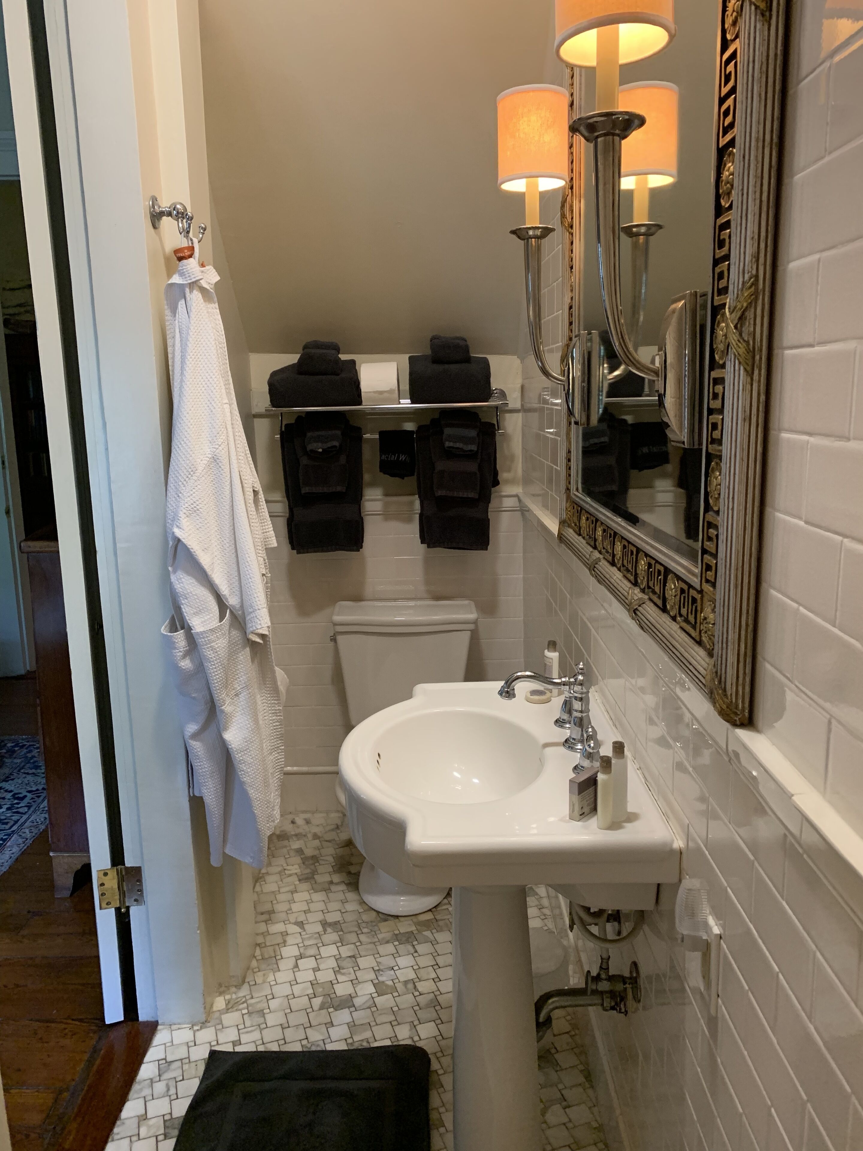 A bathroom with a white sink, a toilet, and a mirror with a decorative frame. There are towels hanging on a rack and a small shelf with toiletries.
