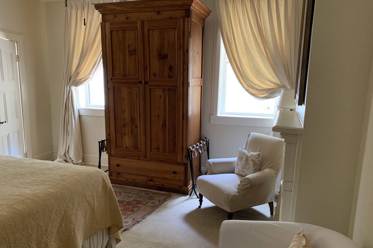 Quarto com guarda-roupa de madeira grande, poltrona branca, cama com colcha bege e janela com cortinas.
