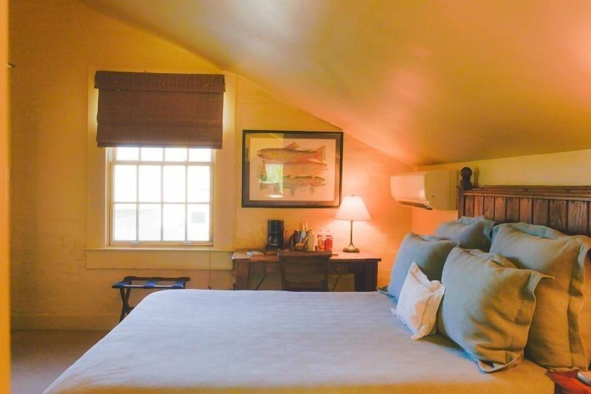 Quarto com cama, escrivaninha, luminária, quadro de um peixe emoldurado e janela com cortina.