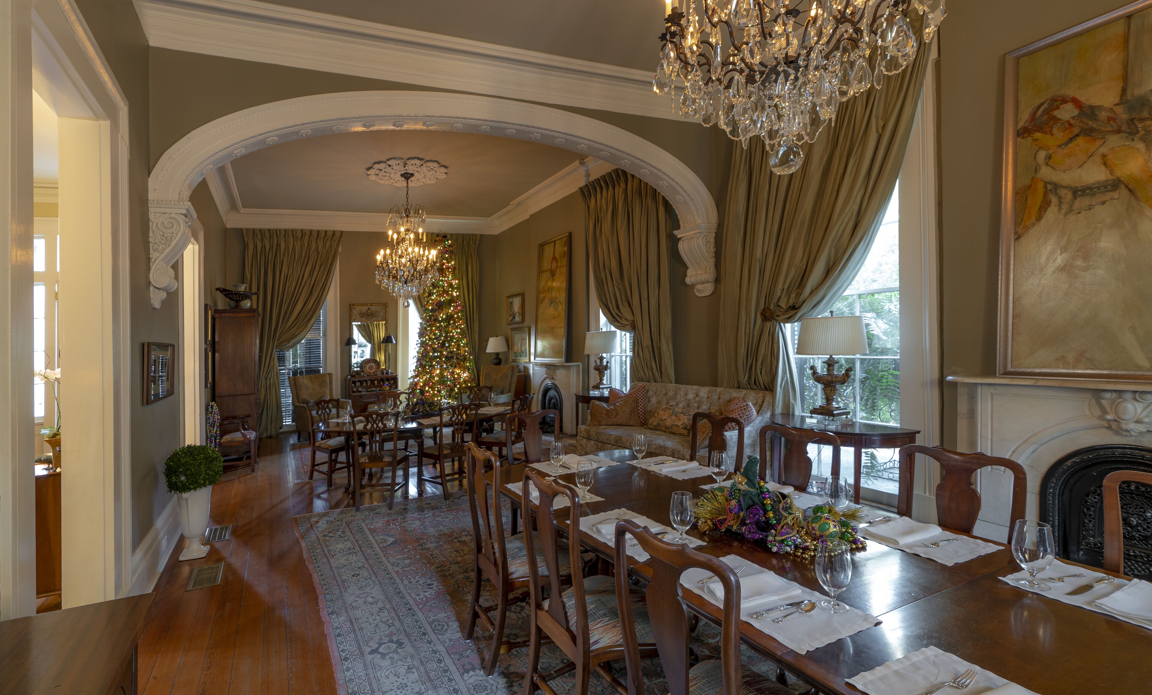 A dining area with a large wooden table set for six, surrounded by chairs. There is a chandelier above the table and a fireplace to the right.