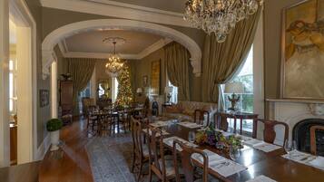 A dining area with a large wooden table set for six, surrounded by chairs. There is a chandelier above the table and a fireplace to the right.