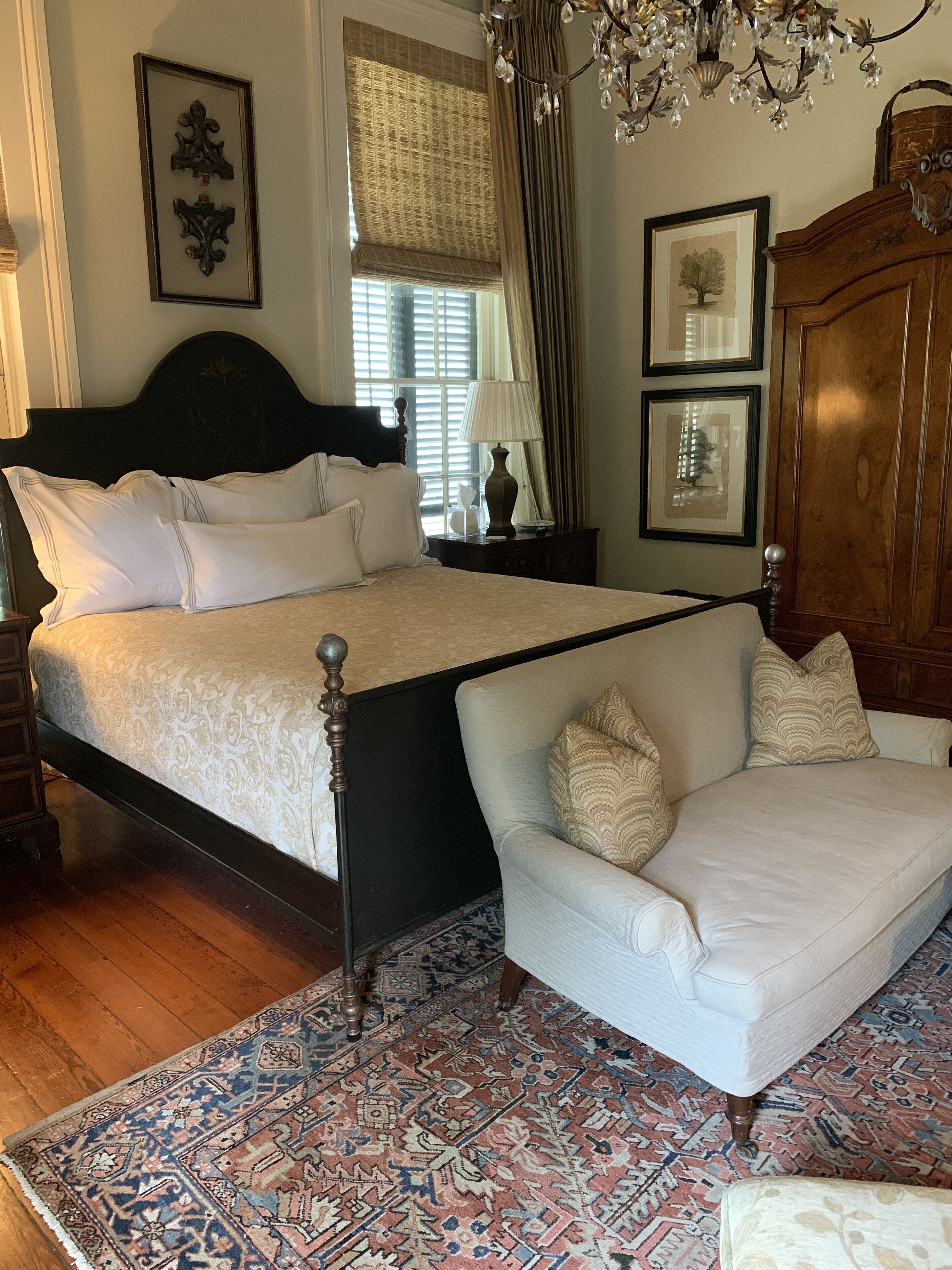 A bedroom with a bed, a chair, a chandelier, and a wooden wardrobe.