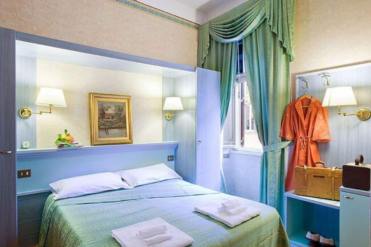 A hotel room with a bed, two bedside lamps, a window with curtains, a painting on the wall, and a robe hanging on the door.