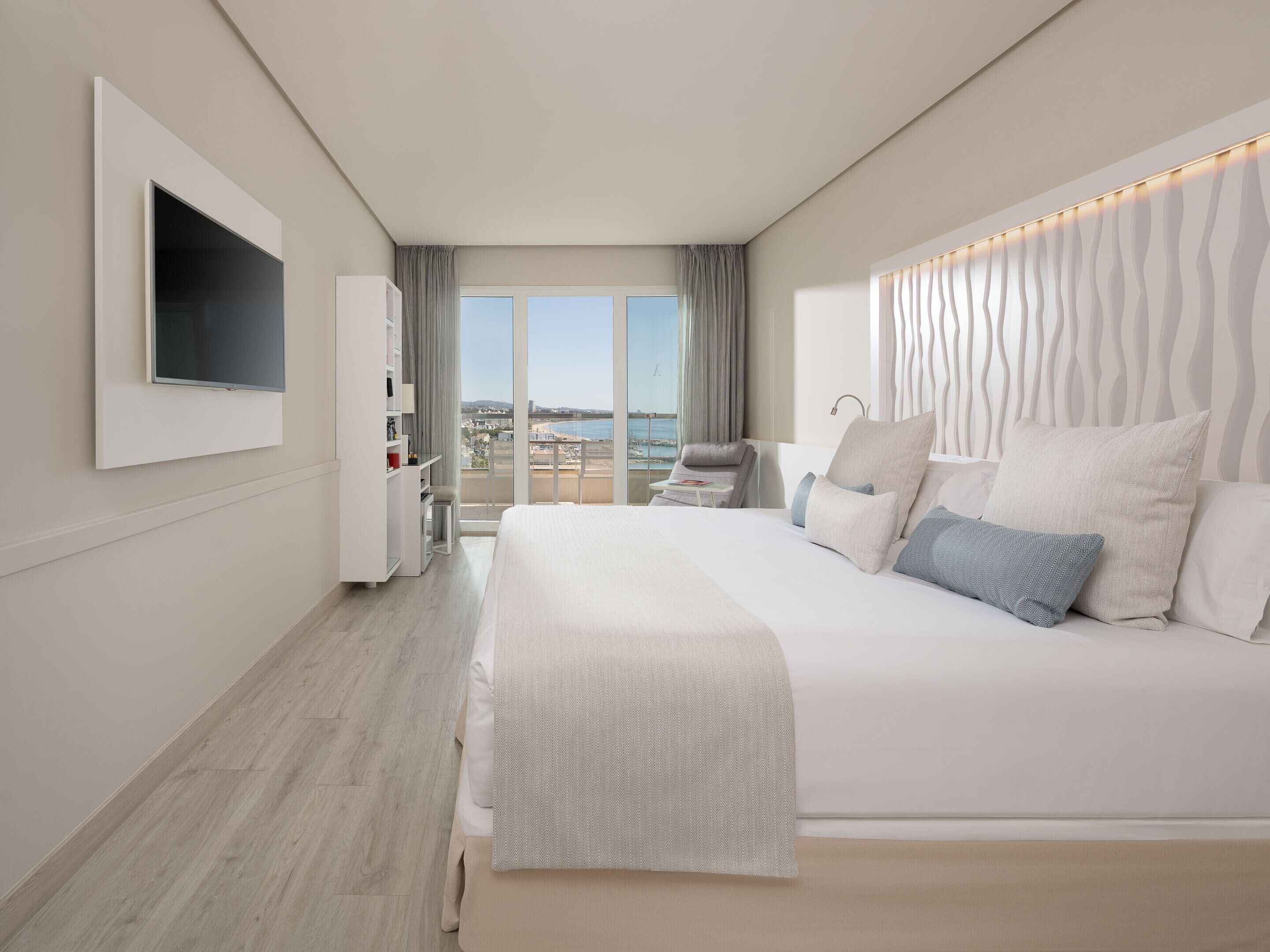 Una habitación de hotel moderna con una cama grande, un televisor montado en la pared, un balcón con vistas al mar y una mesita de noche ordenada.