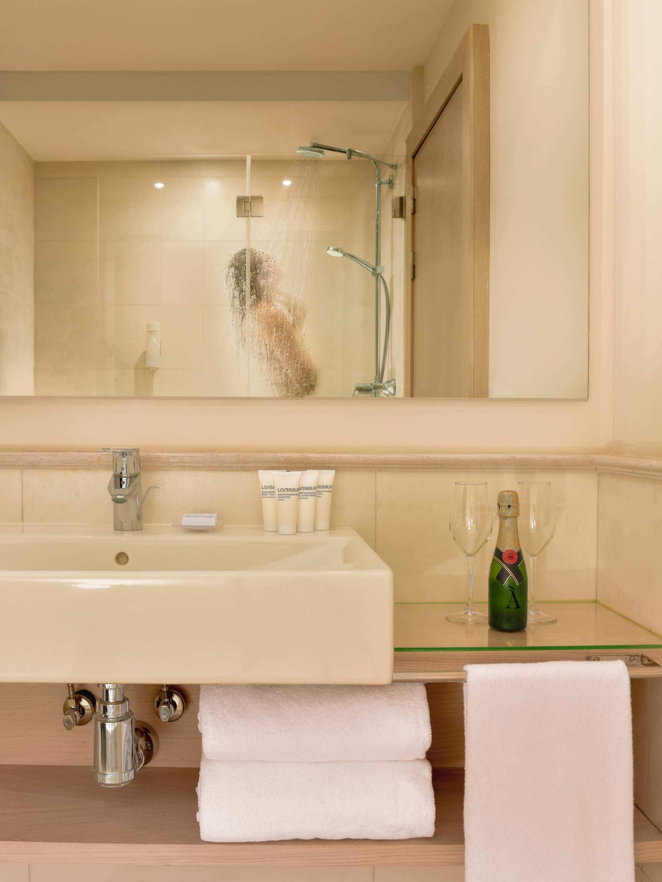 Une salle de bain avec un grand miroir, un lavabo, une douche et une bouteille de champagne posée sur le comptoir.